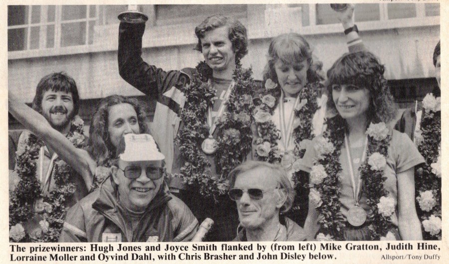 1982 London Marathon winners Hugh Jones and Joyce Smith standing  on a podium and raising their trophies. Both have garlands of greenery and flowers round their necks.