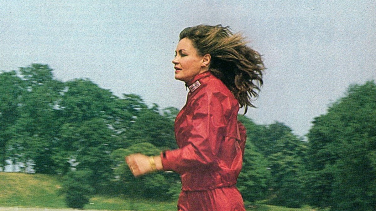 A side view of Leslie Watson running on a track in a red training suit. Her long brown hair is blowing in the wind.