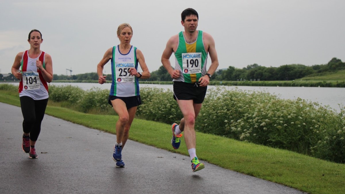 Three runners, a man and two women, competing in the Notts 10 mile road race. They are running on a tarmac path with Holme Pierrepont rowing lake in the background.