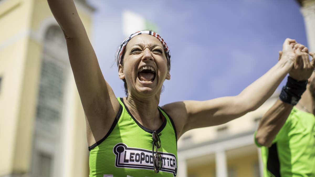 A woman in a green vest shouts in celebration and holds her arms aloft as she finishes the 100km Del Passatore race. Women's participation in ultrarunning.