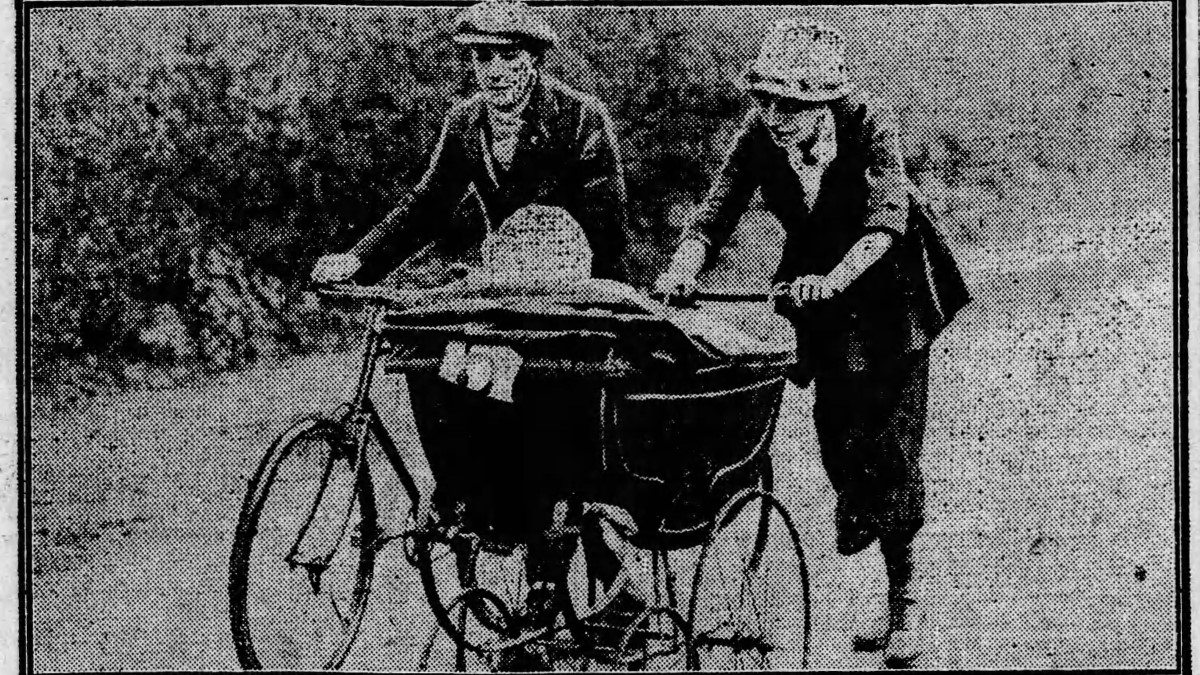 Lily Charlotte Groom winner of the London to Brighton Mothers' Marathon is shown pushing her baby in big black pram accompanied by a woman on a bicycle. Groom is wearing a skirt, jacket and blouse and a light coloured hat.