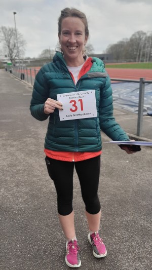 Irisih ultrarunner Aoife Ni Mhaoileoin before the start of the Crawley 24 Hour Track ultramarathon. She is facing the camera and smiling and holding her race number in front of her with her right hand. She is wearing a green padded jacket, black capris and pink running shoes.