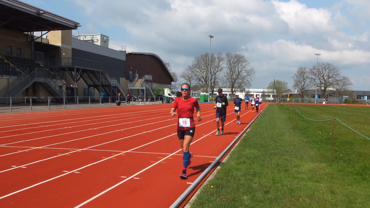 Several runners on the track at the K2 Stadium during the Crawley 24 Hour Track Race. They are running towards us. Eloise Eccles is the first runner. She is wearing a red top and black shorts. The sun is shining and there are clouds in the skyl