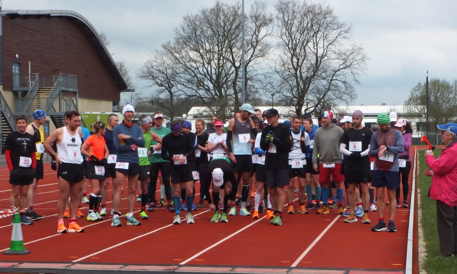 A group of runners wait at the start of the Crawley track ultras. Pam Story stands on the right holding the starter's horn. The timing mat is a few metres in front of the runners.