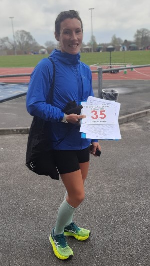 British ultrarunner Sophie Power before the Crawley 24 Hour Track race. Sophie is smiling and looking at the camera. She's wearing a blue jacket, shorts and green running shoes. She's holding her race number in her right hand.