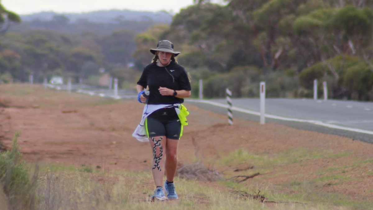 Nikki Love ultrarunner over 50 on her run across Australia. She is wearing a wide-brimmed hat and a dark shirt and dark shorts. She is running on rough ground with the road to her left.