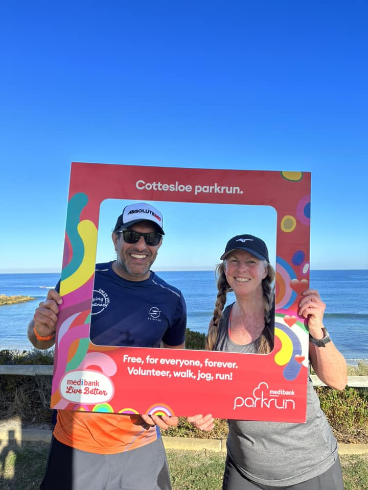 Sharif Owadally and Nikki Love looking through the Cottesloe parkrun photo frame. The sea and blue cloudless sky are in the background.