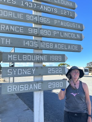 Ultrarunner Nikki Love stands next to a sign with lots of boards showing the distances to different places  - Sydney is 2522 kilometres
