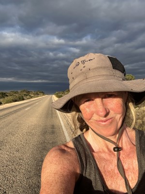 A selfie of Nikki Love on her run across Australia. She is wearing a broad brimmed hat. The road stretches out behind her and there is a dark cloudy sky,