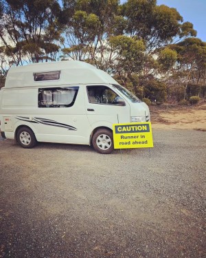 The white van used by Nikki Love for her run across Australia. It is viewed from the side, It has a sleeping compartment on top. In front of the van, there's a yellow sign saying Caution, Runner in Road.