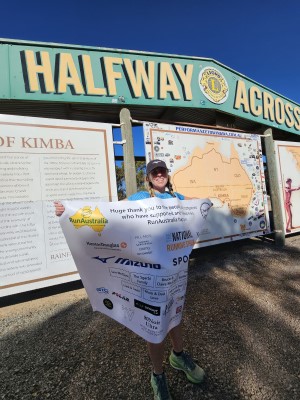 Nikki Love ultrarunner over 50 on her Perth to Sydney run. Nikki stands in front of a large sign saying halfway across. She is holding a banner listing her sponsors.