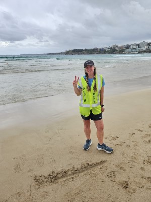 Nikki Love ultrarunner at the end of her 4000km Perth to Sydney run at Bondi Beach. Nikki stands on the sand near the water's edge. She is wearing her yellow hi-vis jacket with the words Perth and Sydney on the front. She holds her right hand in front of her in a victory sign.