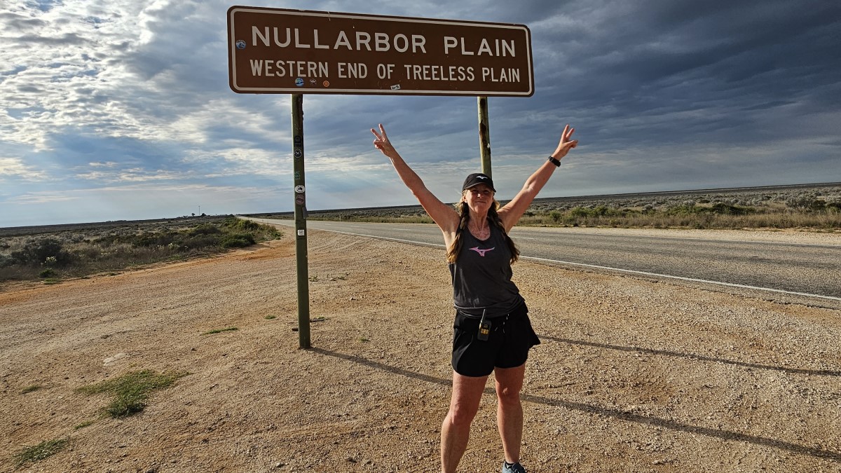 Ultrarunner Nikki Love stands with her arms outstretched above her head in front of a sign saying Nullarbor Plain during her run across Australia