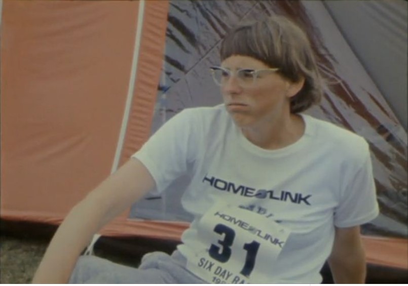 Ultrarunner Eleanor Adams is sitting down and resting during the Charles Rowell 6 Day Race in Nottingham. She wears a white t-shirt with the word "Homelink" and a race number.