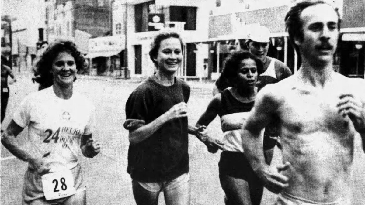 Grainy newspaper photograph of Lorna Richey, Mary Hanudel and Louanne Oberly competing in the Port Clinton Marathon, Ohio in 1984. Richey is to the left of a group of five runners. She is wearing light coloured shorts and t-shirt with race number 28 pinned on it. Hanudel is next to her wearing light shorts and a dark sweatshirt with rolled up sleeves. She does not have a race number. Next to her is Louanne Oberly who is partly obscured by a man racing in front of her with no race shirt and his race number pinned to his shorts.