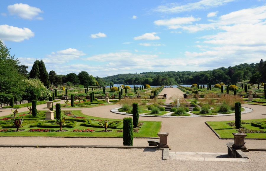 The Italianate Gardens at Trentham Gardens. There are gravel paths and formal borders with grass and plants. A path goes straight ahead to a large round border and then continues down to a lake in the distance. Beyond the lake there are woods.