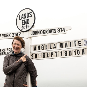 Angela White aka the Running Granny stands in front of a Land's End signpost which has her name on one sign and under it  28th Sept 18D 10H. Angela is smiling and pointing over her left shoulder at the sign.