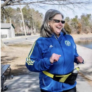 Mary McManus runner is pictured smiling and wearing a blue running jacket and waist belt with a bottle. There are trees and a lake in the background