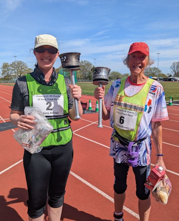 Vicky Barnes and ultra walker Sandra Brown standing on the track at the Crawley K2 arena. They are wearing running clothes and race numbers.