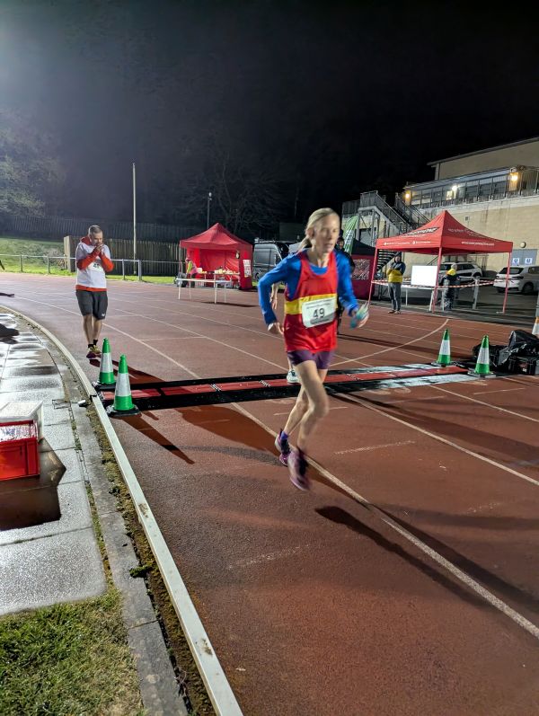 Ultrarunner Sarah Webster is running on a track wearing a red vest over a blue long-sleeved top. It is night-time.